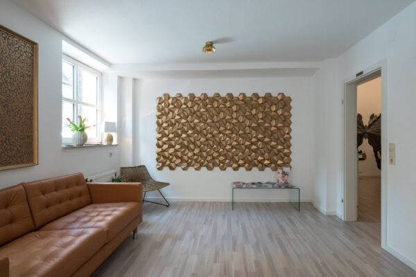 Ein Blick ins Wartezimmer der Praxis Holzfuss Ästhetik. Neben einer Ledercouch sieht man ein goldenes, geometrisches Kunstwerk an der Wand.